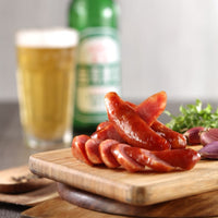 Slicing Taiwanese sausages on a wooden board with beer bottles and glasses in the background, alongside vegetables and onions.