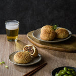 A plate of three sesame seed scones, one on a smaller plate cracked open to reveal a delicious filling flavored with black pepper, rested on a wooden surface next to a glass of beer, with chopsticks and a bowl of burrata in the field of view.