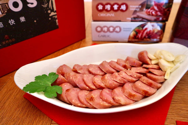 A plate of sliced flying fish egg sausage is neatly arranged with parsley and some garlic cloves on the side. In the background, a promotional box displays Taiwanese delicacies such as famous Penghu products.