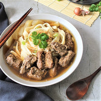 A bowl of noodle soup with sliced pork and cilantro, this soup is a classic Chinese recipe with wooden chopsticks resting on the rim of the bowl, a wooden spoon resting on a nearby gray surface, and garlic and a small cushion visible in the background.