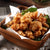 A paper-lined wooden bowl holds a serving of crispy fried chicken nuggets, the meat cash-yellow in color and adorned with fresh basil leaves reminiscent of the popular Taiwanese night markets, the bowl resting on a gray cloth surface.