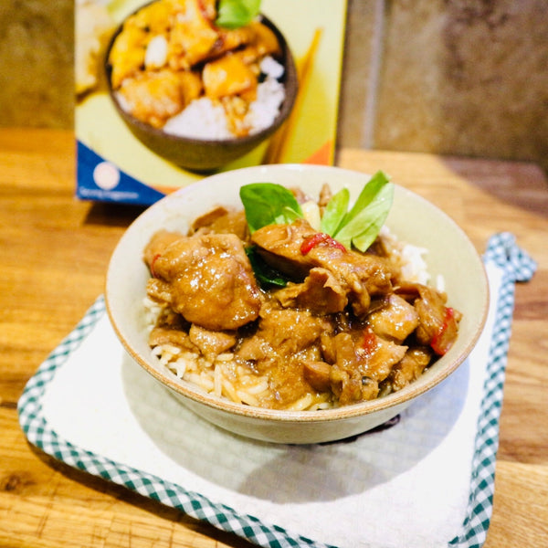 The wooden table is covered with a white cloth with green checkered edges, a bowl of rice is placed on top of a delicious Taiwanese three cups of chicken, a dish containing slices of chicken and vegetables in soy sauce and adorned with green herbs, and in the background there is a box with an image of the meal.