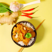 A bowl of rice topped with a delicious Taiwanese three-cup curry chicken adorned with basil leaves, the setup includes ginger root, garlic, red chili peppers, and a white spoon on a yellow background, with the faint scent of soy sauce in the air and chopsticks resting next to the bowl.