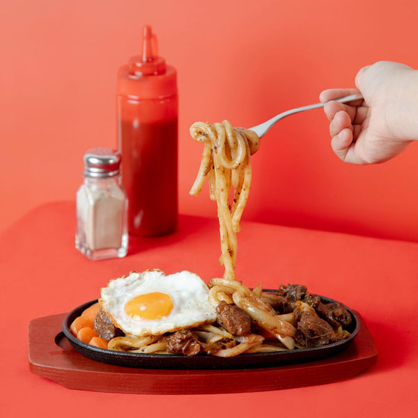 One hand places a forkful of pasta on a plate with black pepper beef udon noodles on top. In the background are ketchup bottles and salt shakers, all against a red background.