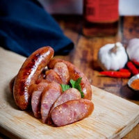 Close-up of sliced Taiwanese sausage on a wooden chopping board. Nearby are garlic, red chili peppers and a spoonful of orange seasoning. A blue cloth and a red bottle are blurred in the background. Enjoying this dish with flying fish eggs will add extra flavor.